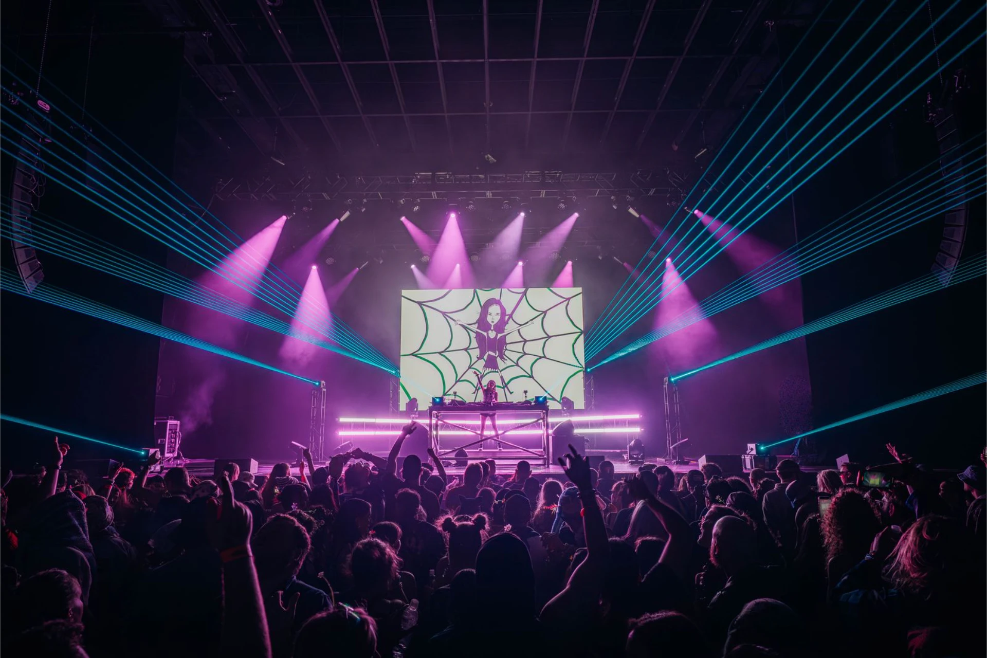 A giant green zombie head with an exposed brain towers over a festival stage as red fireworks and blue lasers light up the night above a packed crowd